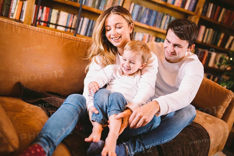 Christmas family portrait. Young Caucasian family in identical clothes having fun on sofa in living room with large library of books and Christmas tree in evening. Happy New Year and Merry Christmas