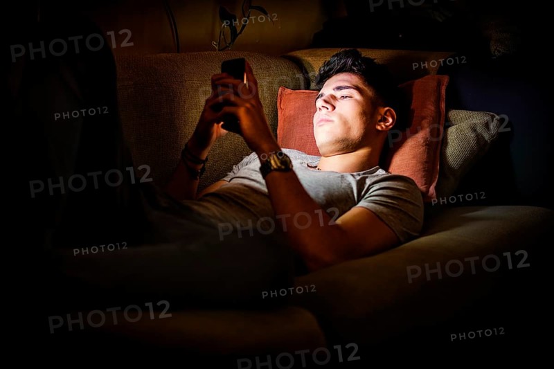 Attractive Young Man Lying on Sofa at Night and Illuminated by Light from Screen of Cell Phone at Home in Living Room