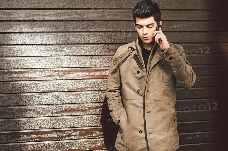 Portrait of a model young mulatto male Turkish Mediterranean race brunet in coat uses hand phone technology businessman on the street in sunny weather against the background of a wooden wall