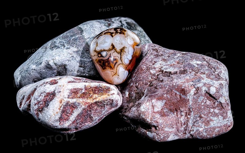 A smooth, polished agate gemstone with intricate banding patterns rests on a group of textured, multicolored rocks against a black background. The image highlights the contrast between the stones glossy surface and the rough texture of the rocks, Odessa, Ukraine, Europe