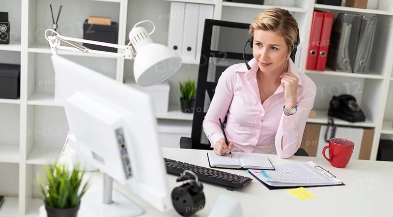 A young girl in headphones sits at a table in the office, holds a pencil in her hand and works with a notebook and a computer