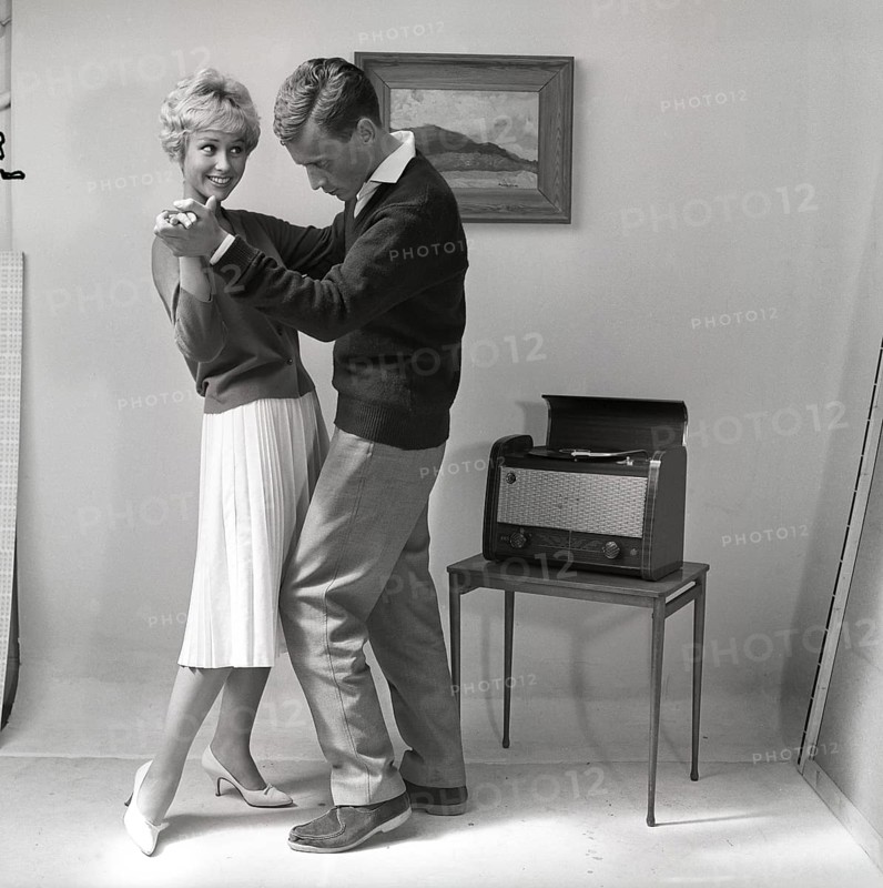 Learning to dance in the 1950s. A young copule is dancing in a photographers studio. The young woman seems to be the one who knows how to dance. Her boyfriend does not seem to know the steps and is struggling with the dance moves. To create the mood of an at home environment a painting is put on the wall and a combined radio and gramophone is visible. Sweden 1959. Kristoffersson ref CG36