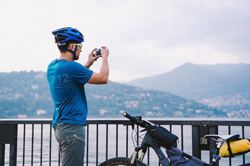 Cyclist taking pictures with smart phone. Caucasian man in bike helmet taking photo of landscape. Outdoor activity. Tourist makes photo of Lake Como in Italy. travel people lifestyle
