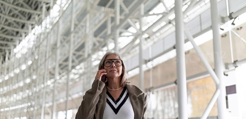 A middle-aged businesswoman with glasses is engaged in a phone conversation while strolling through a bright and spacious airport terminal. Her confident demeanor suggests professionalism and purpose