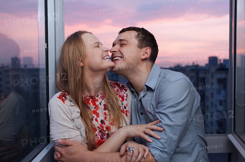 Playful young couple embracing and laughing on the balcony at a high floor during sunset