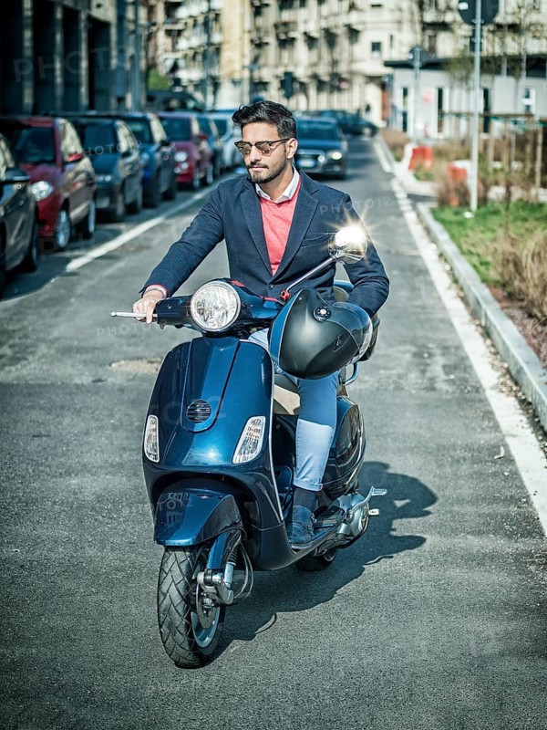 Young stylish man in suit and sunglasses driving scooter on empty road in sunny city