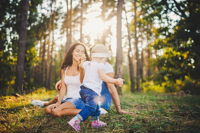 Young parents are taught to walk a daughter of one year on nature in a park on a lawn. The first steps of the child's hand with his mother and father