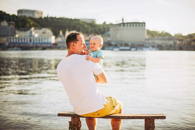 Subject recreation father and little son. Young caucasian dad sits on wooden bench overlooking the town Kiev and holding river of the Dnieper, hugging a toddler baby on sandy beach on the banks pond