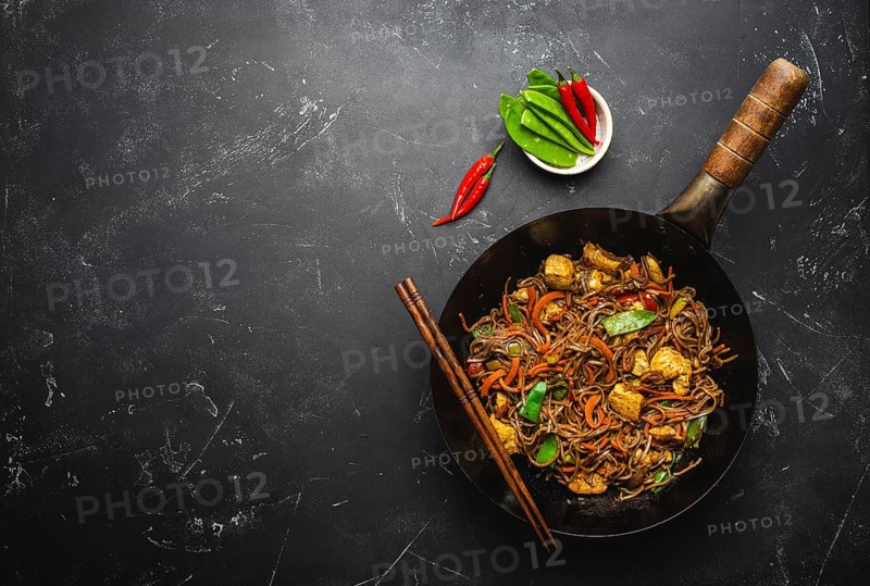 Stir fry soba noodles with chicken, vegetables in old rustic wok pan, chopsticks on black stone background, close up, top view. Traditional asian/thai meal, space for text