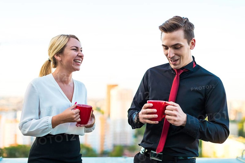 Couple of caucasian businessmen, young attractive brunette man in dark suit, red tie and black shirt, and pretty blonde woman in white blouse and black skirt stand on the roof with red cups of coffe, having a conversation, guy is shy blured city on background