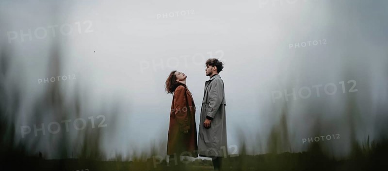 A couple standing in a field, each looking upwards, dressed in long coats with a sense of connection
