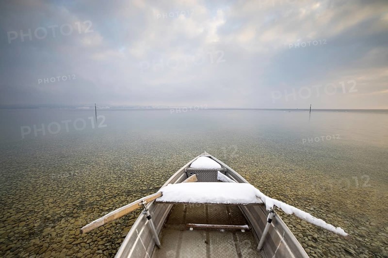 A snow-covered boat on a calm lake with pebbles on the shore under a cloudy sky, Strandbad Hörnle, Constance, Lake Constance, Baden-Württemberg, Germany, Europe
