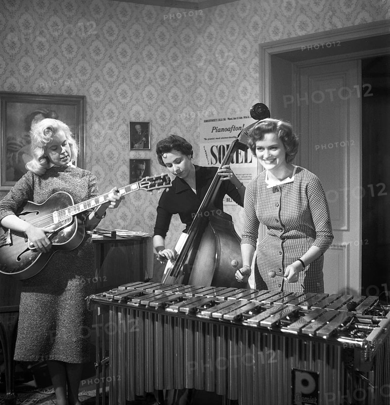 Trio 1956. The photograph, taken in 1956, captures three young women in a band, each immersed in their performance. One strums an electric guitar, her fingers poised over the strings with confidence, while another stands beside a double bass, plucking its deep notes with focus. The third plays a xylophone, her mallets striking the wooden bars in a lively rhythm. The scene blends the bold, emerging sound of mid-20th-century music with an unconventional lineup, showcasing a striking mix of instruments and the women's evident talent against a backdrop of 1950s style. Kristoffersson ref BS4-3
