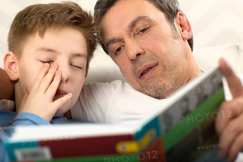 Close-up shot of a father reading a book to the son who is already sleepy and rubbing his eyes