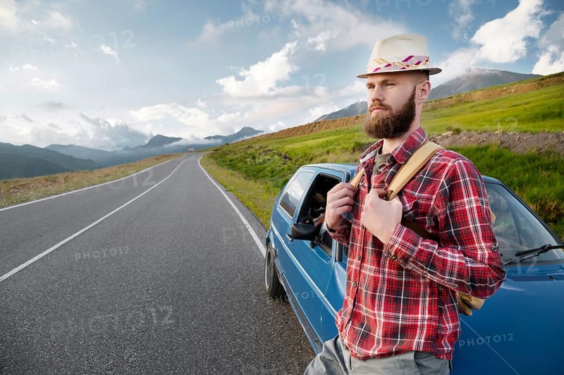 Attractive bearded man in a plaid red shirt and hat with a backpack. Stands next to a retro car on the background of a picturesque road in the mountains at sunset. Easy travel, hitchhiking
