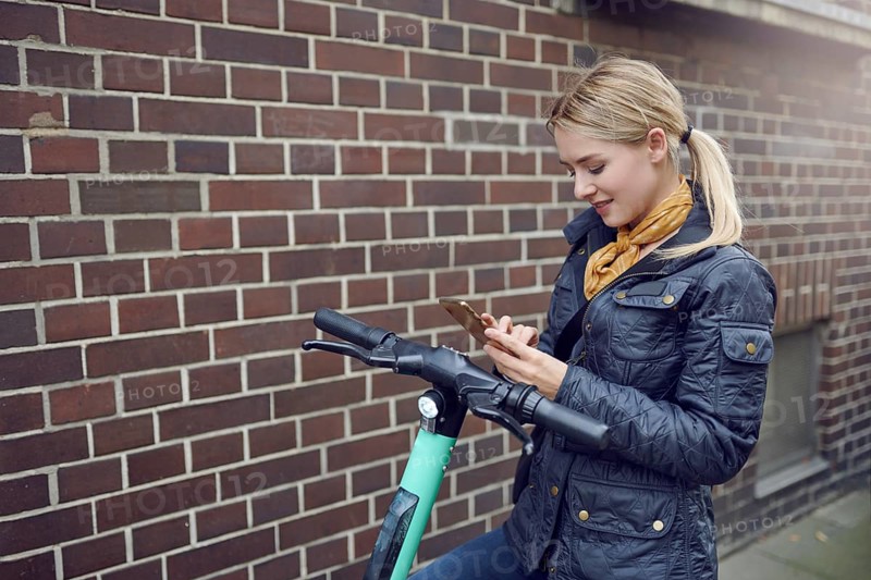 Trendy young blond woman in leather jacket standing at her e-scooter booking a ride, ecological transportation concept with copy space