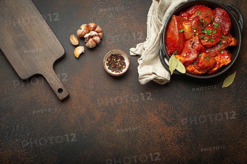 Cooking meat stew. Raw uncooked chopped pieces of meat marinated with seasonings and parsley in black casserole dish top view, cutting board, ingredients on dark rustic background. Space for text, food photography, food photography