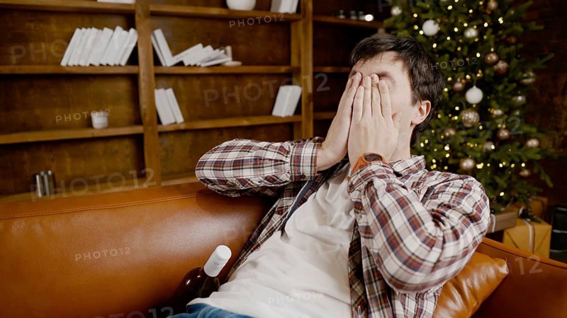 Young male alcoholic requires medical and psychological help. Man addicted alcohol and cannot stay on couch at home on Christmas and New Year Eve. Drunk guy drink wine from bottle near Christmas tree