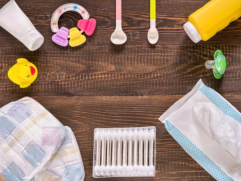 Japanese diapers, wet wipes, baby powder, cream, tither, cotton buds, spoons, soother and duckling on dark brown wooden background with copy space. Flat lay or top view