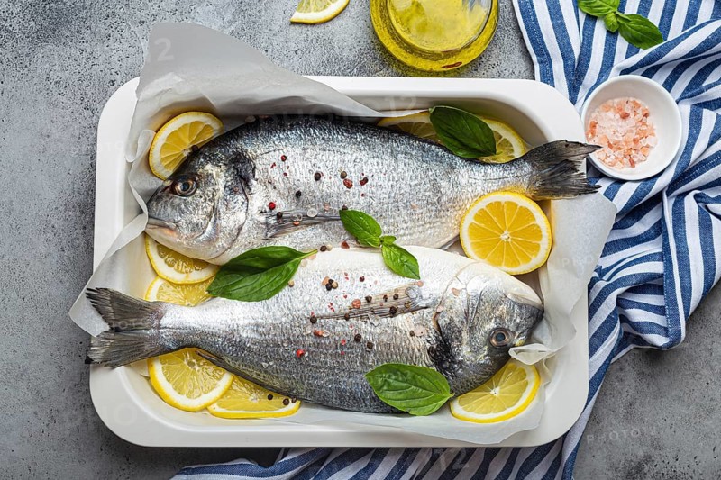 Raw fish dorado in casserole dish with ingredients lemon, fresh basil, bottle of olive oil on wooden cutting board with knife on rustic stone background top view, cooking healthy fish dorado concept, food photography, food photography