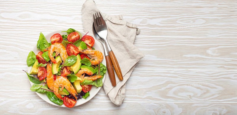 Healthy salad with grilled shrimps, avocado, cherry tomatoes and green leaves on white plate with cutlery on white wooden rustic background top view. Clean eating, nutrition and dieting, copy space, food photography, food photography