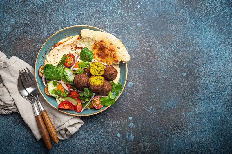 Middle Eastern Arab meal with fried falafel, hummus, vegetables salad with fresh green cilantro and mint leaves, pita bread in ceramic bowl on stone rustic background top view, space for text, food photography