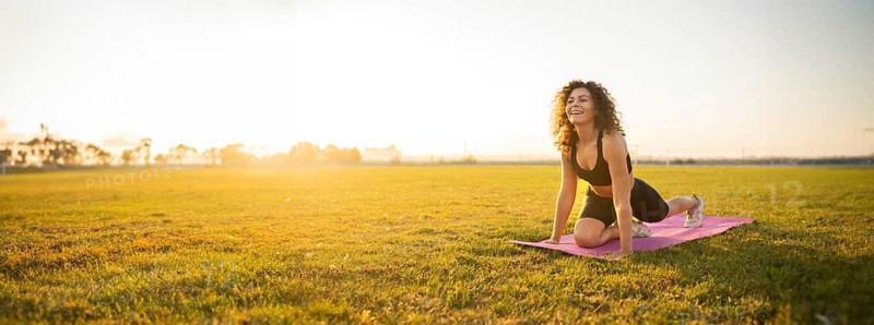 Person smiles while in a yoga pose on a mat, lit by the morning sun, in a field