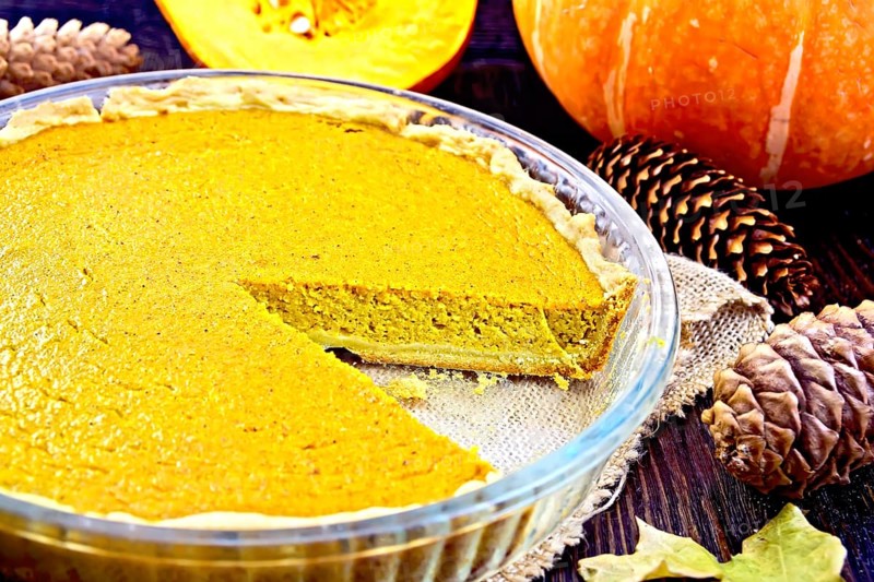 Pumpkin pie in a glass pan on sackcloth, pumpkins, pine cones and maple leaf on the background of dark wood planks