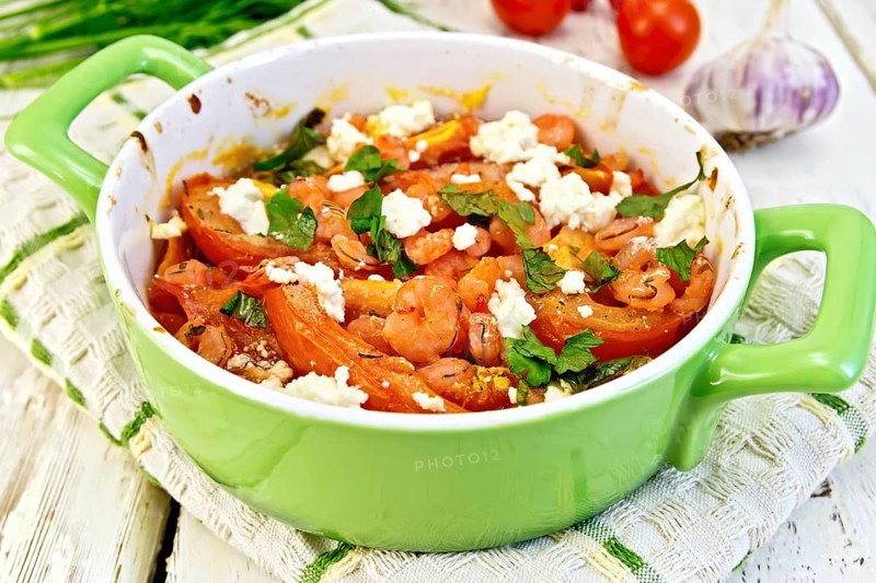 Shrimp and tomatoes baked with feta cheese in a roasting pan on a towel, parsley and garlic on a wooden boards background