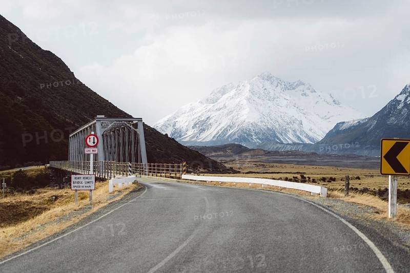 Road leading to a bridge with snow-capped mountains and cloudy sky in the background, Hooker Valley Track, New Zealand, Oceania