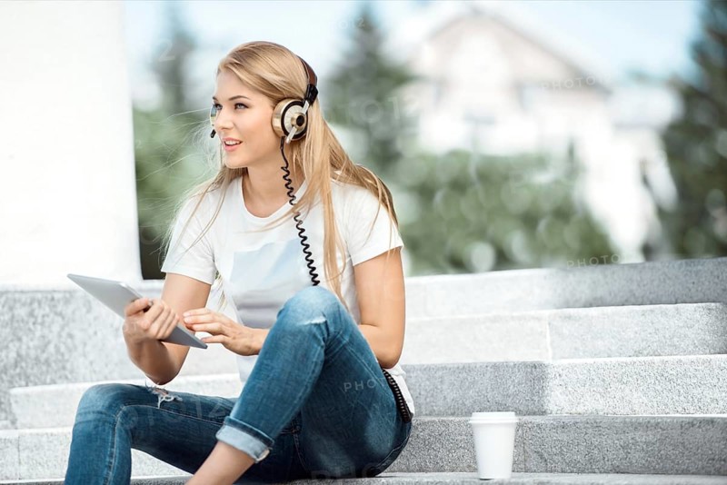 Happy young woman with vintage headphones and a cup of coffee to go, surfing the internet on a tablet PC, listening to music and sitting on a staircase in front of an urban background