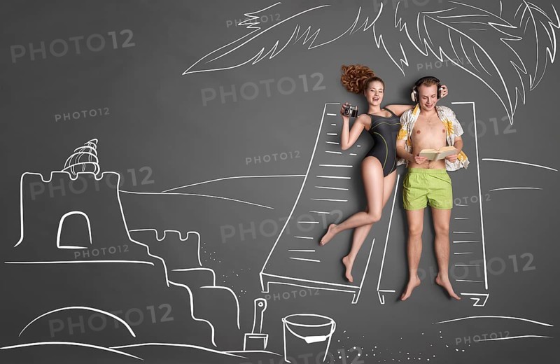 Love story concept of a romantic couple against chalk drawing background. Male lying on sun lounger wearing headphones and reading a book, female taking a picture of a sandcastle