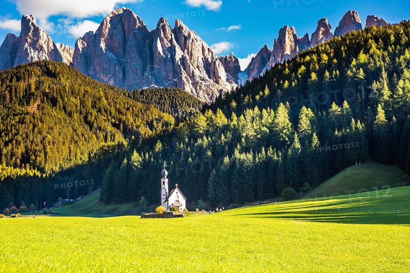 Dolomites, Tirol. Sunny autumn day. The famous symbol of the valley Val di Funes - church of Santa Maddalena. Rocky peaks and forested mountains surrounded by green Alpine meadows