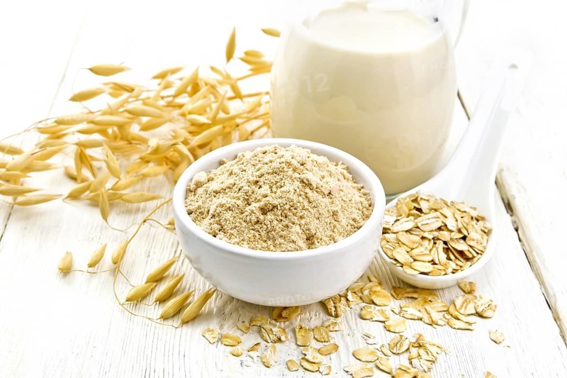 Flour oat in bowl, milk in a jug, oatmeal in spoon, oaten stalks on the background of light wooden board