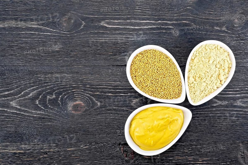 Mustard sauce, seeds and mustard powder in three saucepans on a wooden plank background