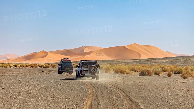 Off-road vehicles drive on a dusty road in front of large sand dunes, With off-road vehicles through the Sahara desert in Algeria