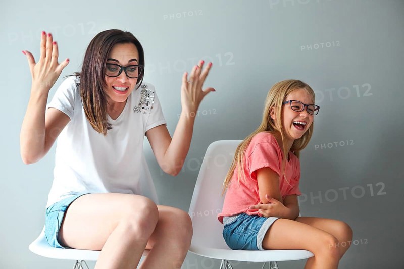 Happy mother and her little daughter sitting on the chairs by the grey wall. Portrait of mother and daughter with eyeglasses