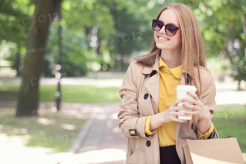 Young fashionable woman taking a coffee break after shopping and walking with a coffee to go in her hand against the backdrop of a green city park