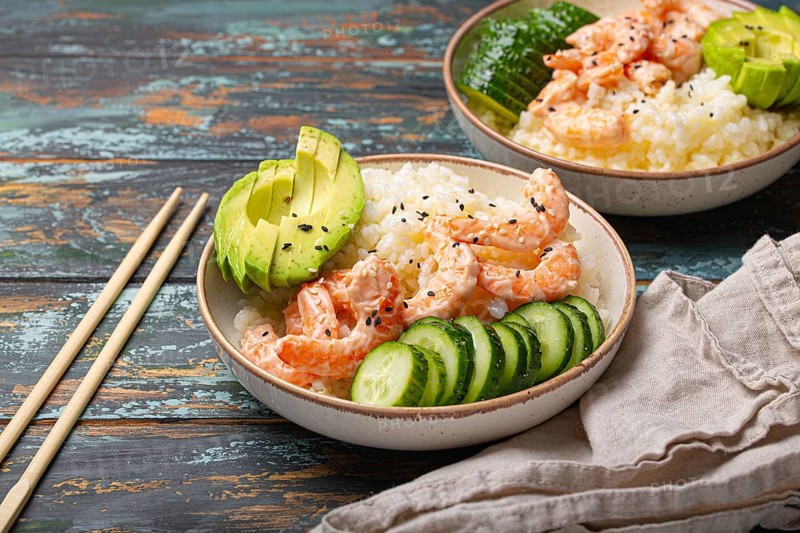 White ceramic bowl with rice, shrimps, avocado, vegetables and sesame seeds and chopsticks on colourful rustic wooden background front view. Healthy asian style poke bowl