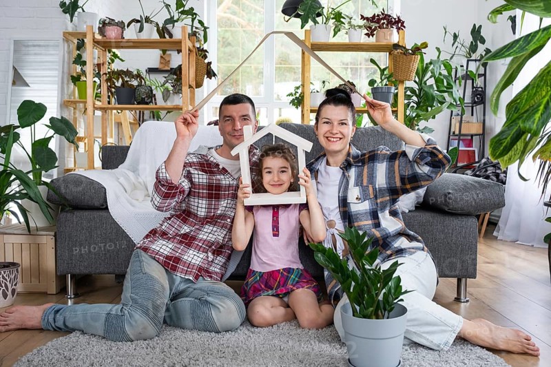 Traditional family with child are enjoying new home, sitting on the sofa and holding the roof. Mortgage, insurance and protection, buying and moving into your own home, green house with potted plant