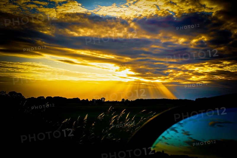 A dramatic sunset with yellow and blue tones, rays of light through clouds and a dark silhouette in the foreground and reflections in a car rear-view mirror, Polder 27798 Hude, Germany, Europe