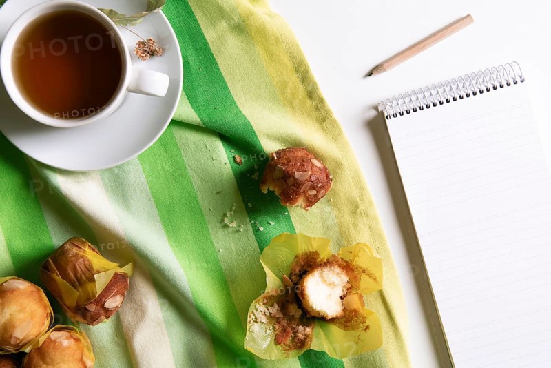 Above view of a breakfast table with a cup of linden tea, muffins on a green and yellow towel and an unwritten notebook, on a white table