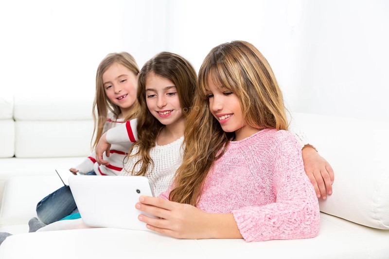 Three kid sister friends girls group playing together with tablet pc on white sofa