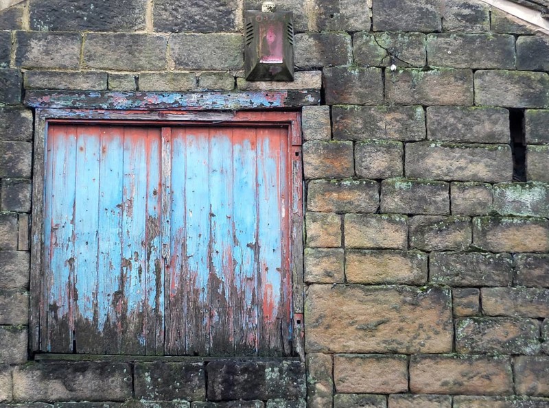 Boarded window with decaying blue painted shutters with rotting wood in a derelict abandoned house with stone walls