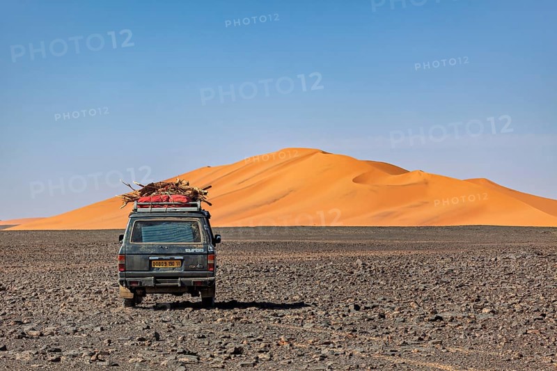 Off-road vehicle leads through a rocky plain in front of large sand dunes under a blue sky, By off-road vehicle through the Sahara desert in Algeria