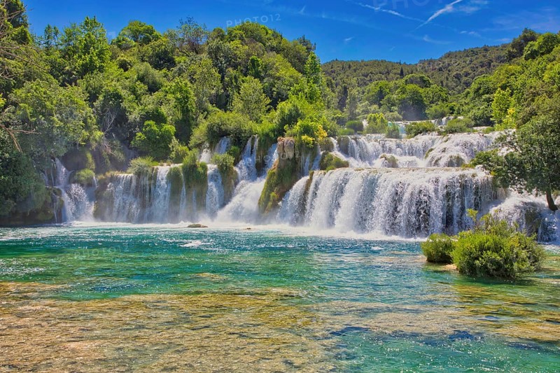 Waterfalls flow into an emerald green river, surrounded by dense forest and a bright blue sky, Krka National Park Lozovac Dalmatia Croatia