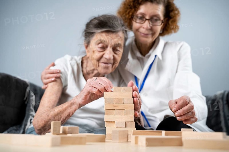 Senior woman practice skills, build wooden blocks, build tower and try not to let it fall, Jenga game. Old patient pull out block, place on top, support doctor during therapy dementia in house
