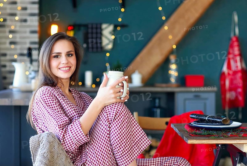 Happy young woman in plaid pajama holding jar smiling at camera in kitchen decorated for Christmas