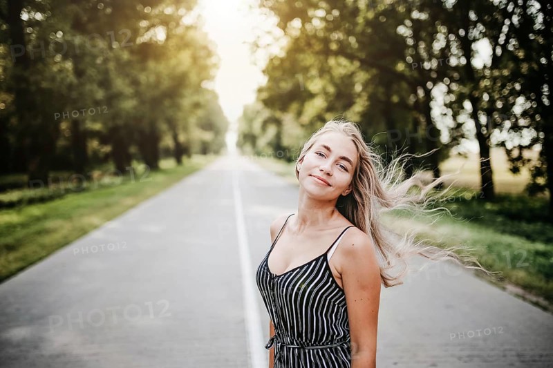 Woman standing on a road surrounded by trees, sunlight filtering through, creating a relaxed summer atmosphere