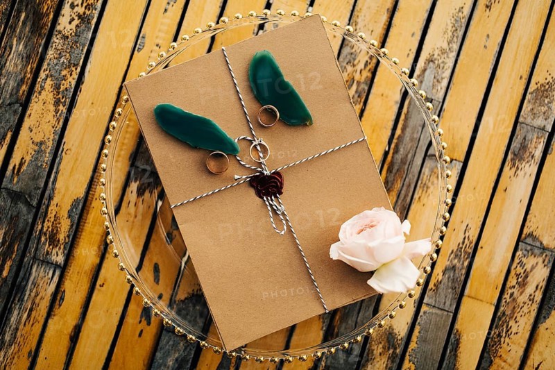 Beautiful brown envelope tied with a rope with rings, a rose and petals lies on a transparent tray on a wooden table.